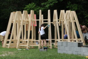 Workers and volunteers assembling the wooden modular frames of the structure in an outdoor setting, demonstrating the self-construction process