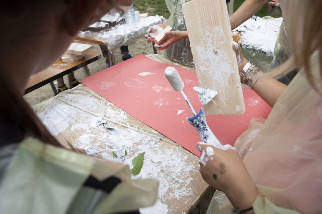 Close-up of children's hands using paint rollers and natural leaves to create white patterns on red fabric during a workshop to create a sense of belonging