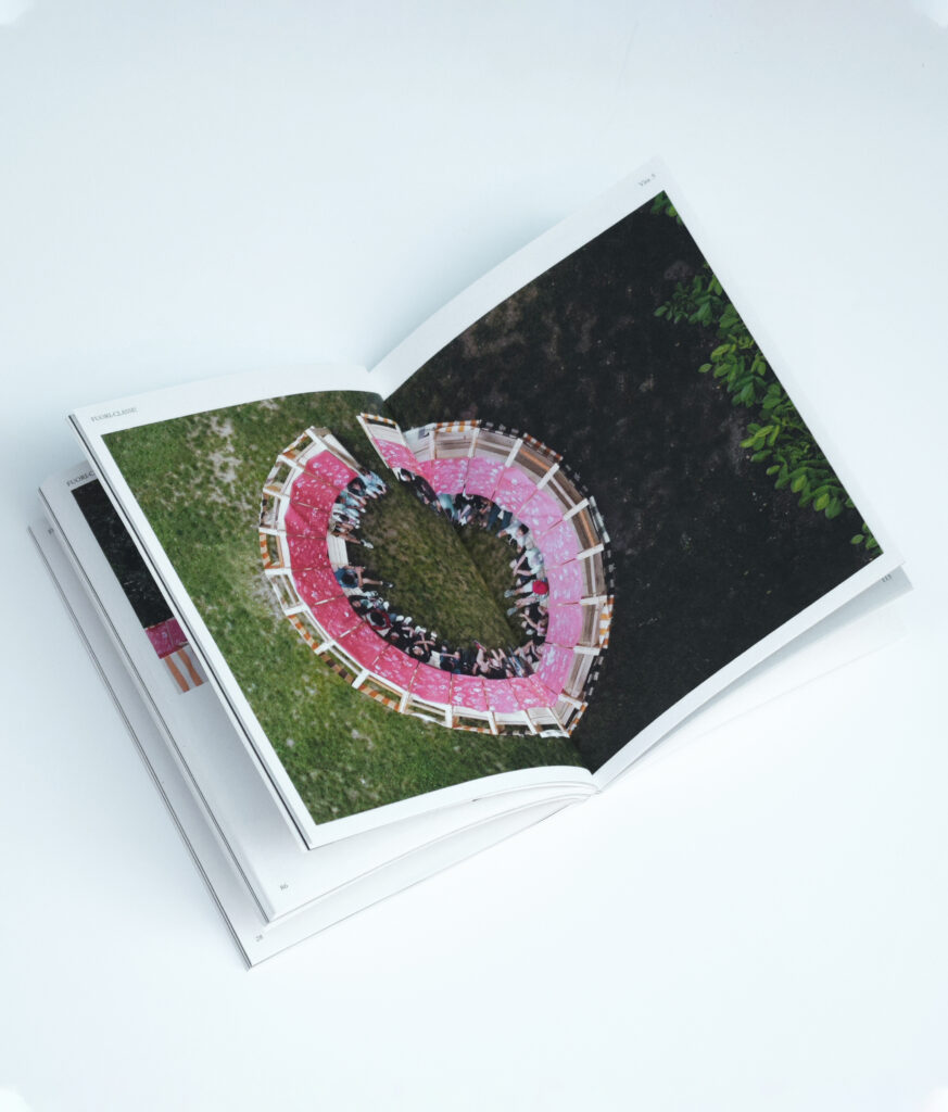An open book featuring a full-page photograph of the completed circular classroom from a high-angle drone perspective, showing pupils and teachers sitting inside the modular wooden structure with red-patterned curtains 
