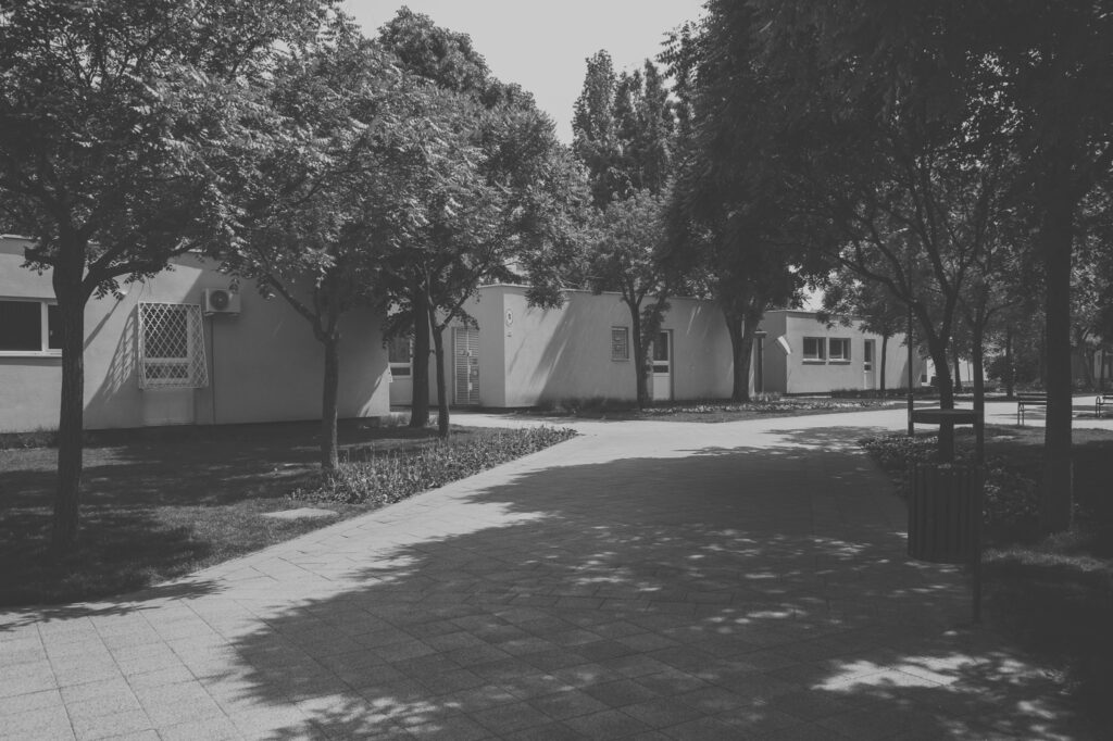 A black and white photograph showing the original flat-roofed, single-story prefabricated kindergarten building and its surrounding tree-lined promenade