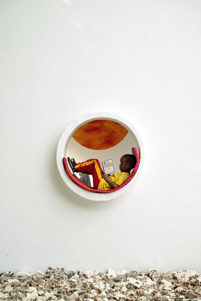 Interior shot of the library highlighting the filtered daylight passing through perforated opaque corrugated panels onto a concrete floor, with a large, colorful circular mural depicting a cityscape on an adjacent white wall.
