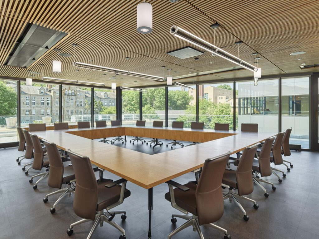 A large, sunlit conference room with a square wooden table, ergonomic leather chairs, and views of the surrounding campus