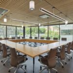 A large, sunlit conference room with a square wooden table, ergonomic leather chairs, and views of the surrounding campus