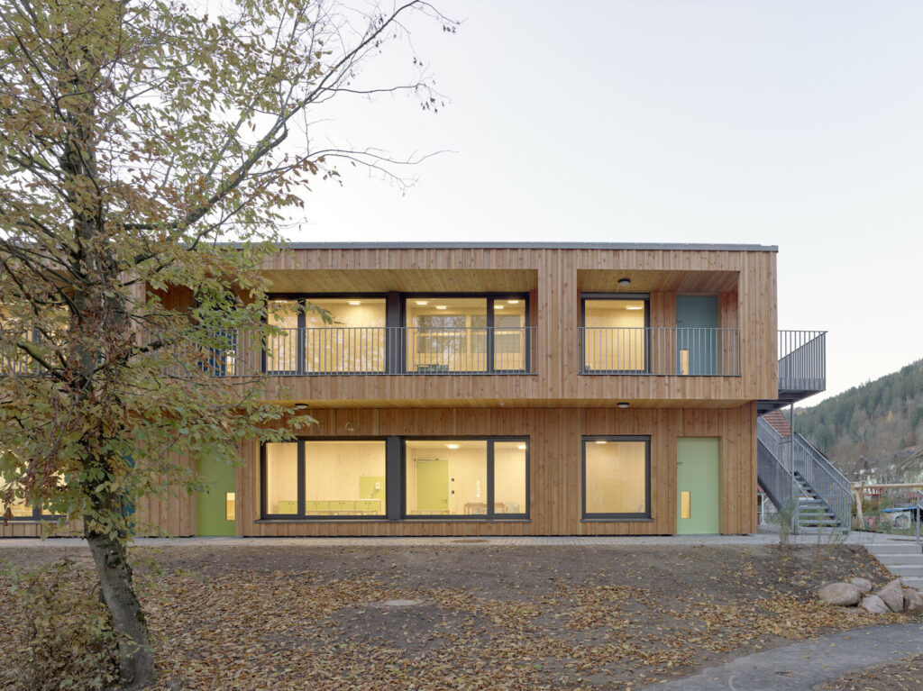 An exterior shot of the kindergarten’s sustainable timber facade, showing large windows and the integration of the building into the sloped site
