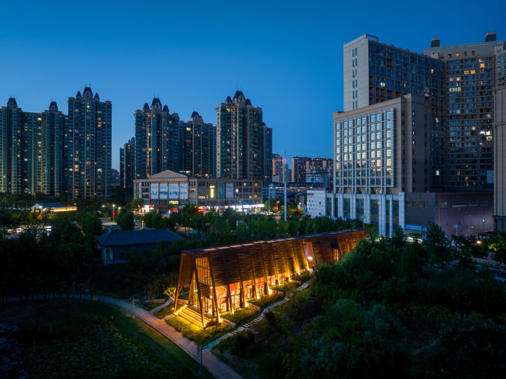 A high-angle view showing the tea house building almost entirely enveloped by a dense canopy of trees in Yangshan Park. The surrounding metropolis of Xinyang is visible on the horizon
