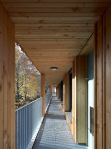 A view looking down the length of the external access gallery, showcasing the wooden structural pillars and metal floor grating