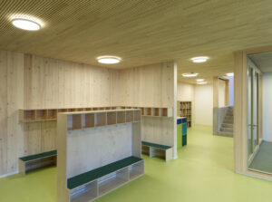 Interior view of a cloakroom with custom-built solid wood storage cubbies, benches, and a bright green floor under circular ceiling lights