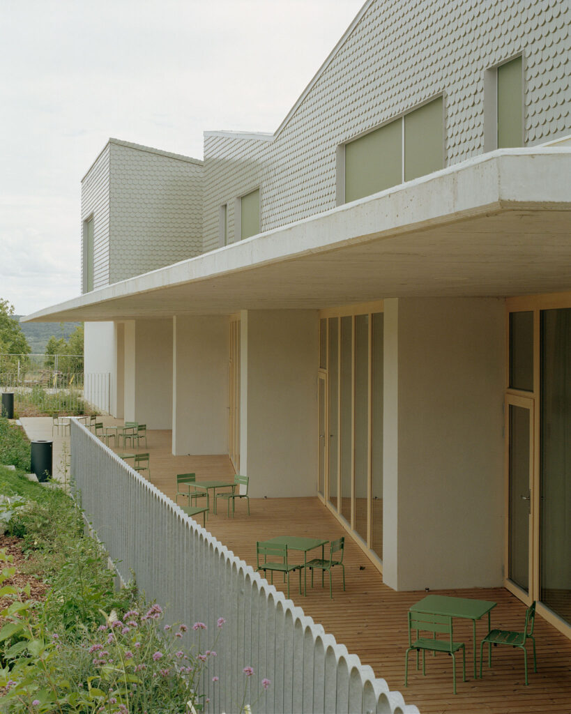 A wooden outdoor terrace at Children’s Grove with green tables and chairs, shaded by a concrete overhang, overlooking the playground