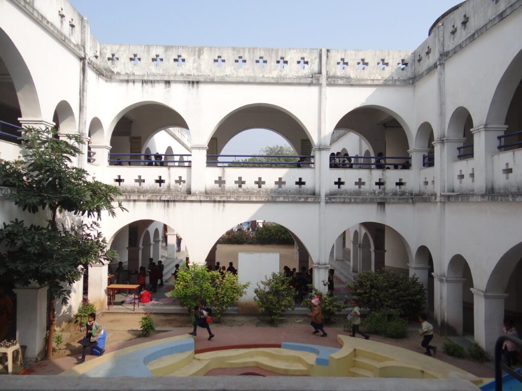A view of white arched corridors surrounding a central courtyard with children playing and a colourful patterned floor at the Nadiad school
