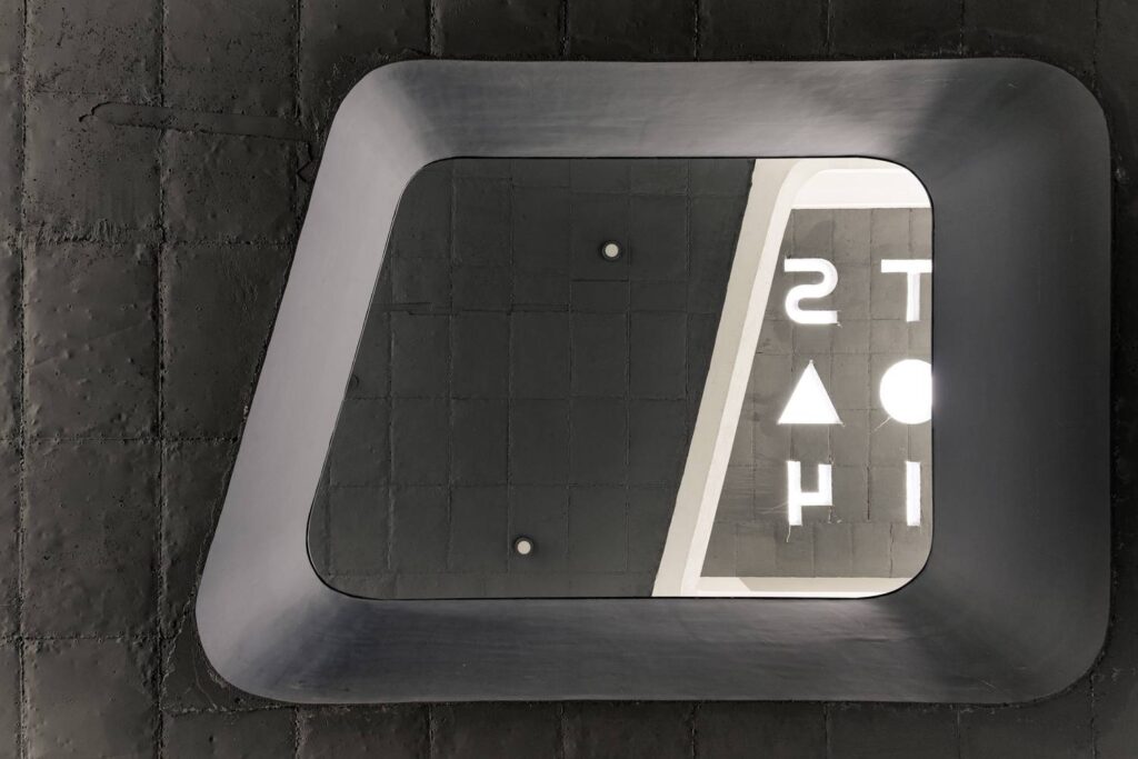 Looking up at a rectangular ceiling opening within a black textured surface, featuring alphabet-shaped cutouts that allow light to filter through for shadow play