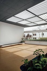A sunlit semi-outdoor terrace featuring a white slatted pergola roof that casts geometric patterns on the floor, intended as a learning medium for children