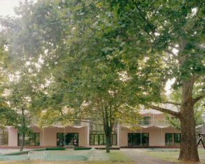 Wide shot from the courtyard showing the two-story building blocks with upper-level galleries and staircases leading down to the play area under large trees