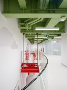An elevated interior walkway with a glass railing and a green industrial ceiling with exposed pipes. A red steel staircase and mountain wall graphics are visible.