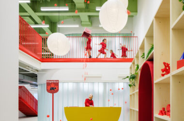 A wide view of the two-story entrance hall featuring exposed green steel beams, large white globe pendant lights, and a yellow semicircular reception desk.