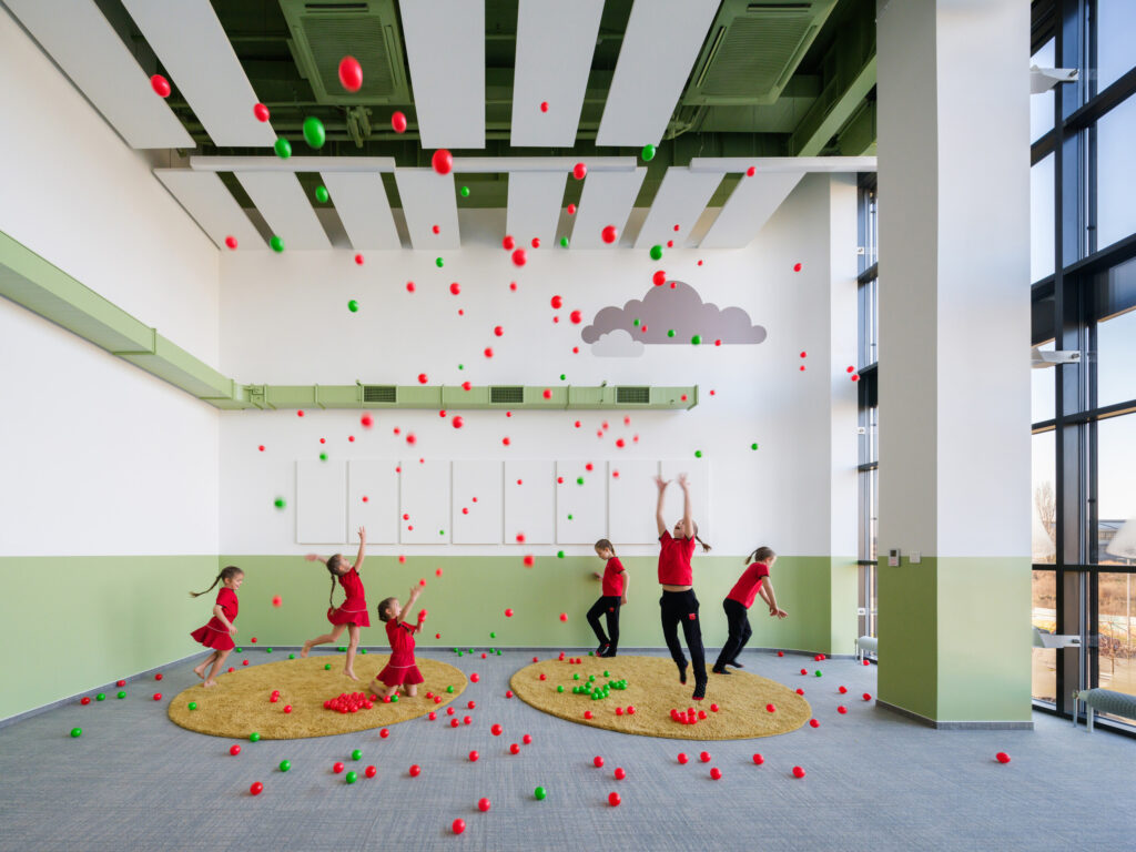 Children playing with red and green balls in a large room with floor-to-ceiling windows, green wall accents, and circular yellow rugs.