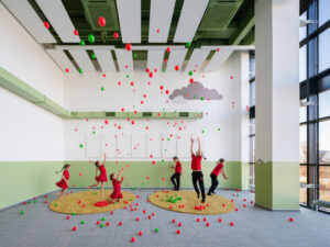 Children playing with red and green balls in a large room with floor-to-ceiling windows, green wall accents, and circular yellow rugs.