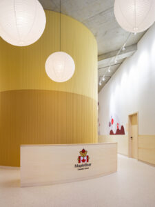A curved wooden reception desk featuring the Maple Bear logo, set against a tall yellow acoustic wall and globe lights.