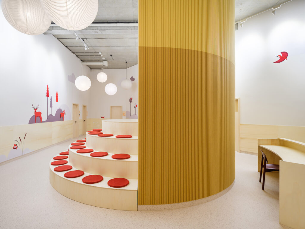 A tiered wooden seating area with red circular cushions built around the base of the yellow cylindrical meeting room.