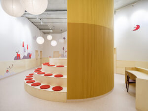 A tiered wooden seating area with red circular cushions built around the base of the yellow cylindrical meeting room.
