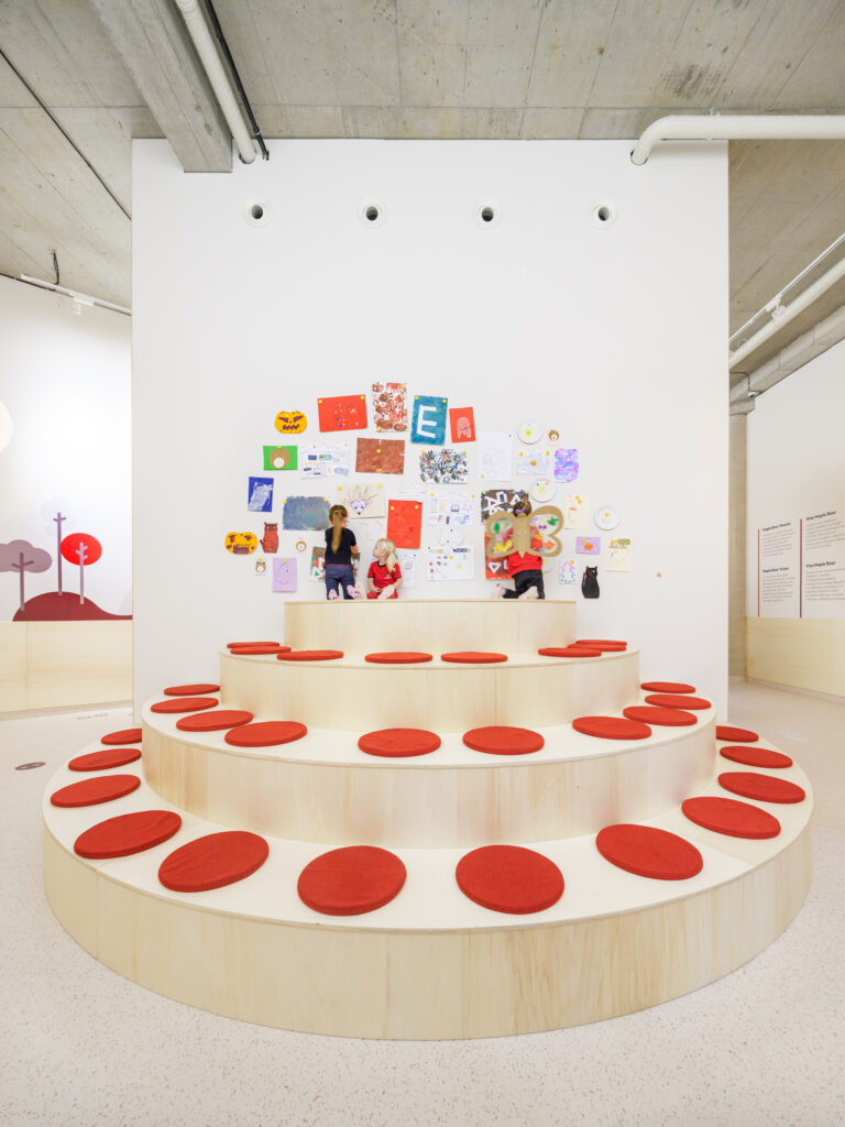 Children sitting on the wooden amphitheater steps below a large wall covered in colorful student drawings and crafts.