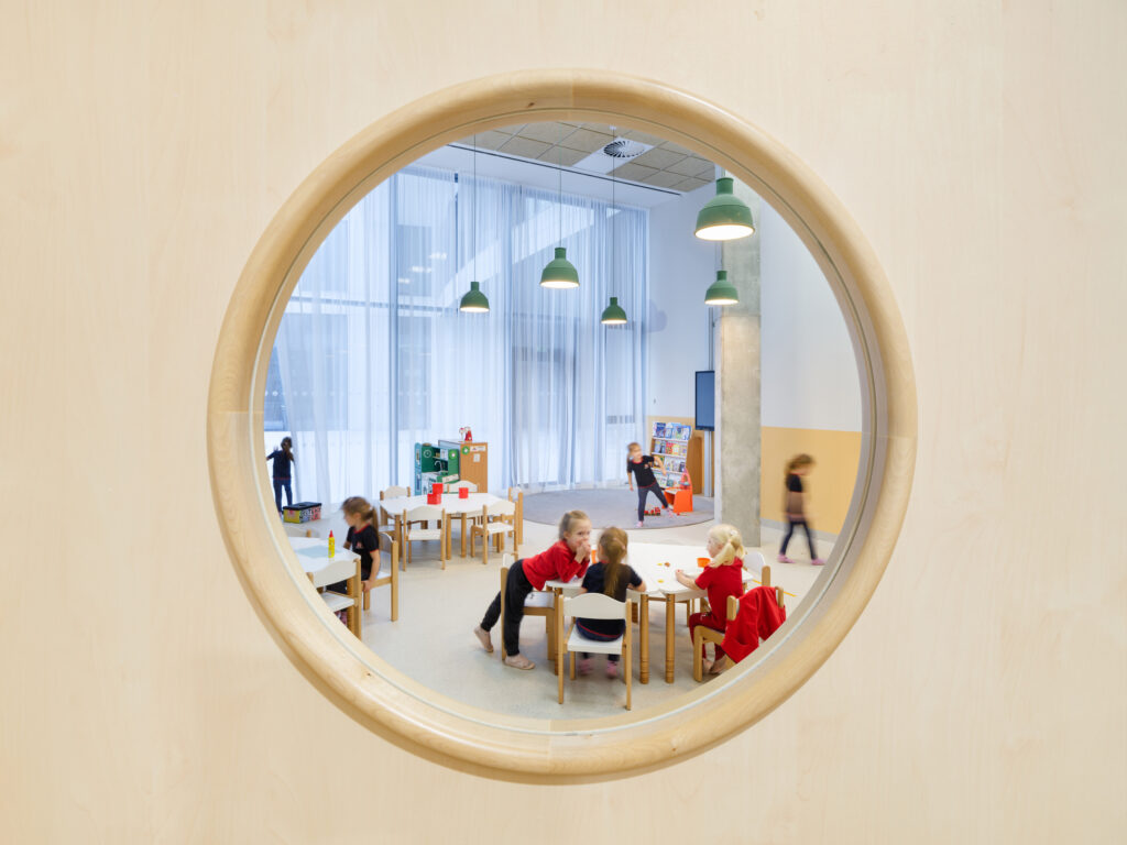 A circular window provides a view of children working at white tables in a sunlit, stimulating environment.