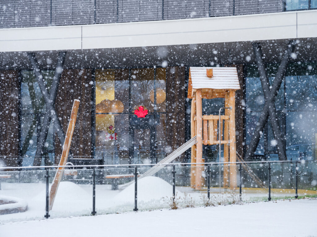 View of the school's exterior and wooden playground structure during a snowfall, with the glowing red Maple Bear logo visible through the glass.