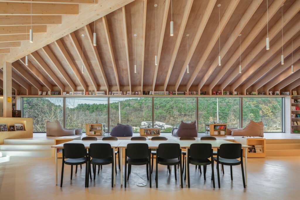 Interior view looking out toward a forest through large windows, with beanbags on a raised wooden platform and a large table in the foreground.