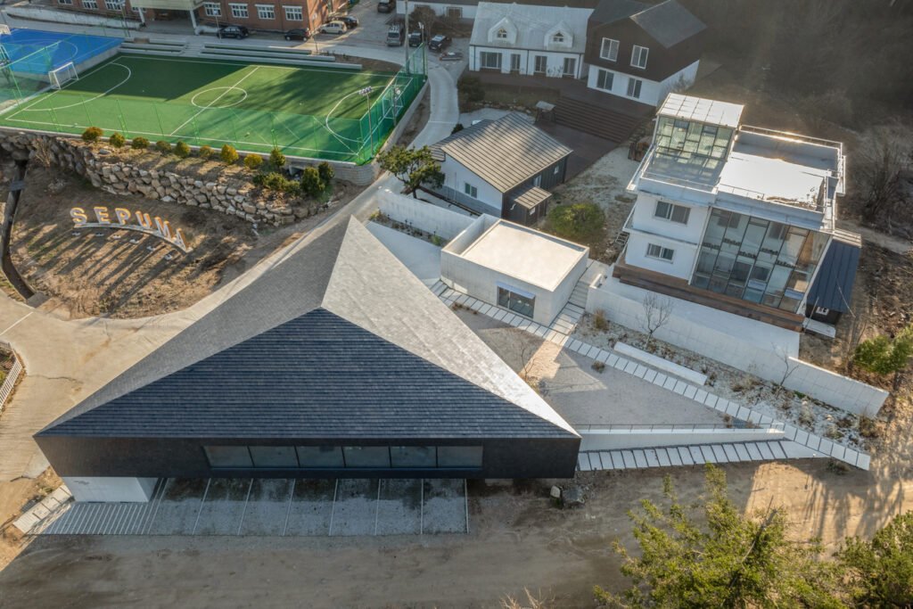 A bird's-eye view of a modern black triangular library building situated on a sloped site next to a sports field in South Korea.