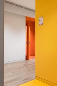 View through a yellow wall toward an orange classroom entrance, demonstrating the use of color as an organizing layer for school navigation.