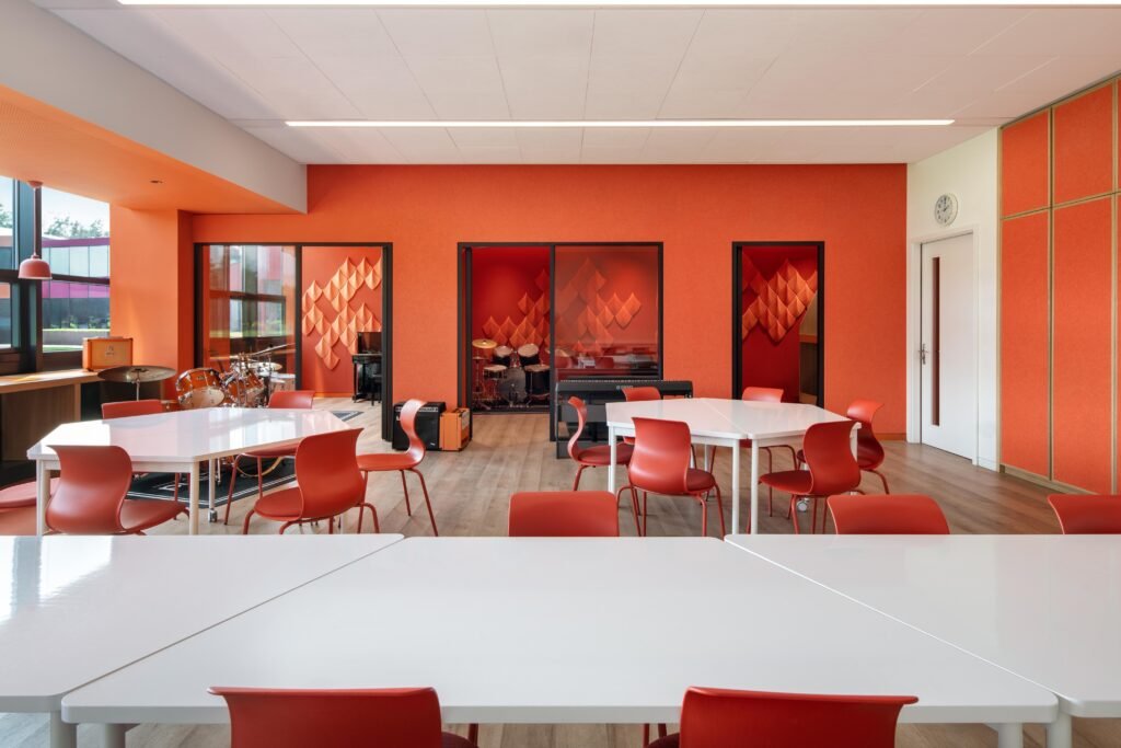 View from a music classroom through glass partitions into adjacent saturated practice rooms, enabling supervision and visual continuity.