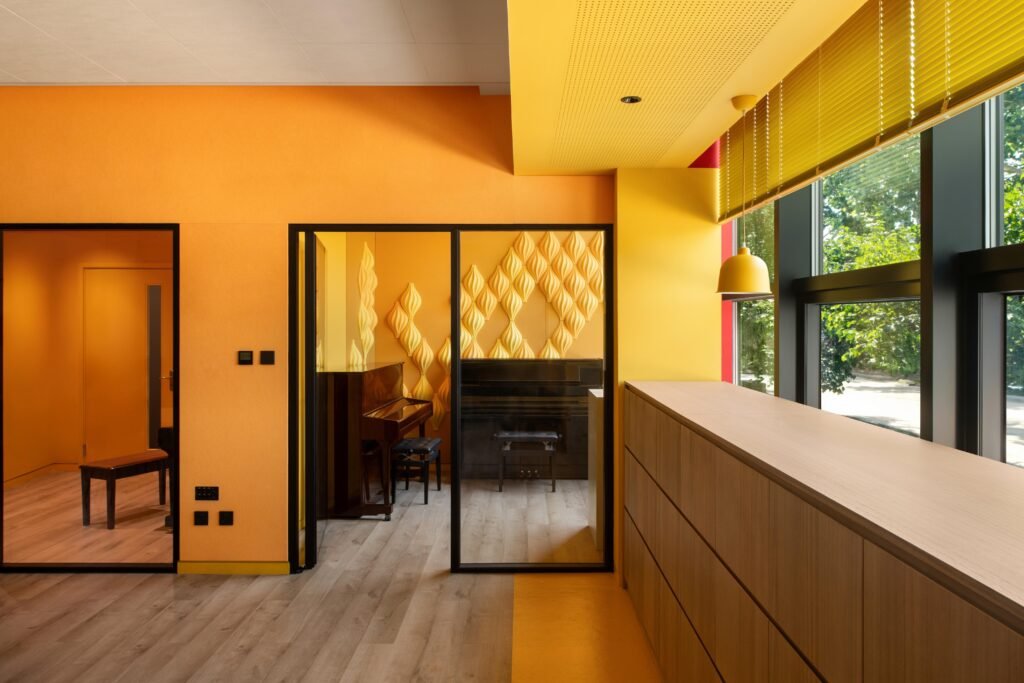 View from a yellow-themed classroom zone toward a saturated practice room featuring glass doors and 3D acoustic wall tiles.