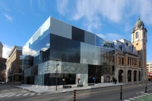 A daytime street-level perspective of La Caserne, showcasing the contemporary checkered glass-clad addition with black, grey, and white panels on the left, seamlessly joining the historic 1912 brick fire station and its domed tower on the right under a clear blue sky