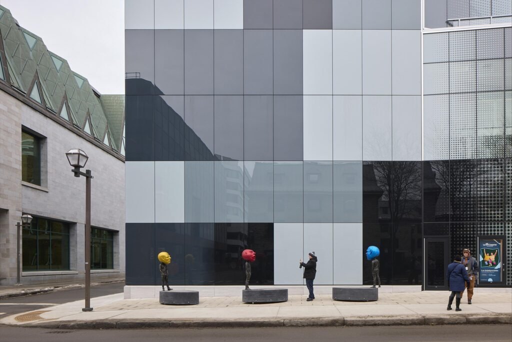 Three whimsical bronze head sculptures painted in vibrant yellow, red, and blue mounted on circular concrete pedestals on the sidewalk in front of La Caserne's checkered glass facade, with a pedestrian pausing to take a photograph