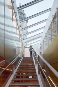 An interior view looking up an open metal staircase under a fully glazed linear skylight roof, where a visitor ascends between reflective stainless steel and gold metallic walls as the historic fire station dome tower is revealed overhead