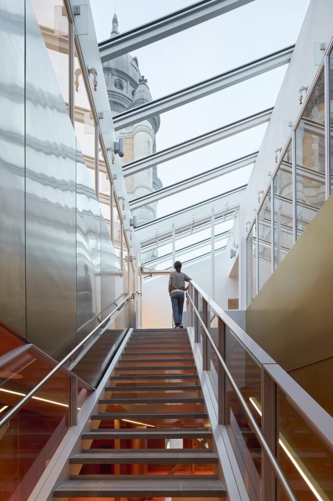An interior view looking up an open metal staircase under a fully glazed linear skylight roof, where a visitor ascends between reflective stainless steel and gold metallic walls as the historic fire station dome tower is revealed overhead