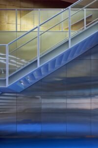 An abstract interior detail highlighting the clean lines of the grey painted steel staircase structure, safety railings, and step profiles reflecting off the wavy mirrored stainless-steel wall cladding