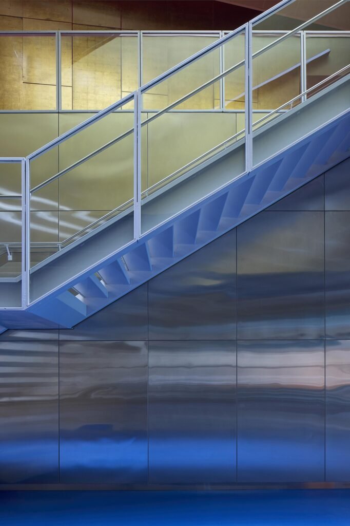 An abstract interior detail highlighting the clean lines of the grey painted steel staircase structure, safety railings, and step profiles reflecting off the wavy mirrored stainless-steel wall cladding