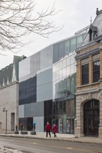 A detail shot of the contemporary glass-clad facade of La Caserne, with its black and grey checkered grid panels reflecting the copper triangular dormers of the neighbouring Musée de la Civilisation