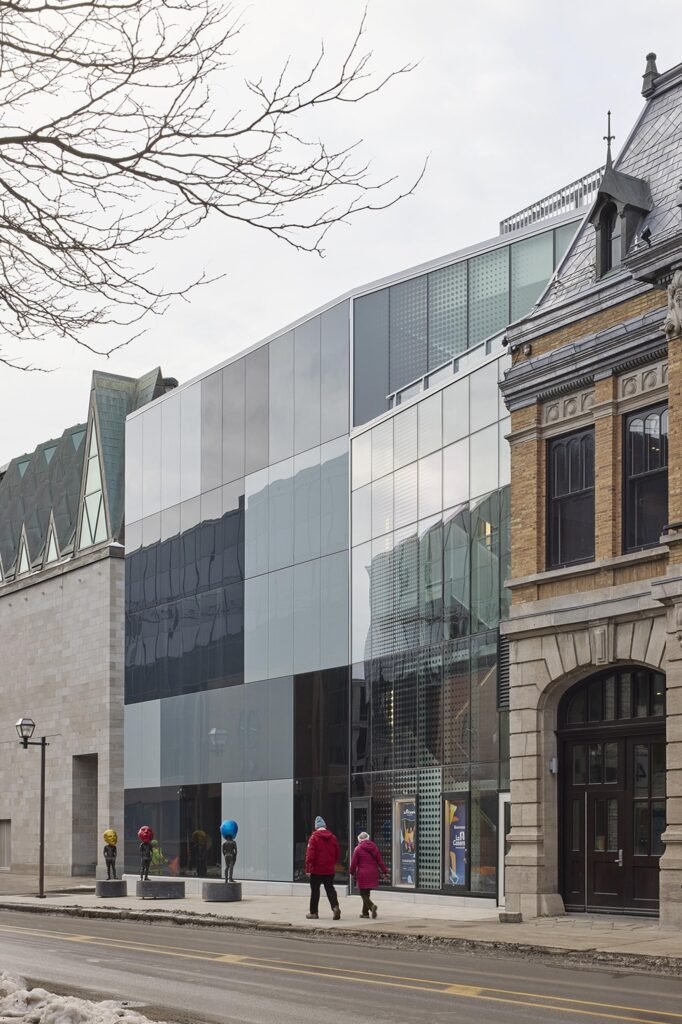 A detail shot of the contemporary glass-clad facade of La Caserne, with its black and grey checkered grid panels reflecting the copper triangular dormers of the neighbouring Musée de la Civilisation