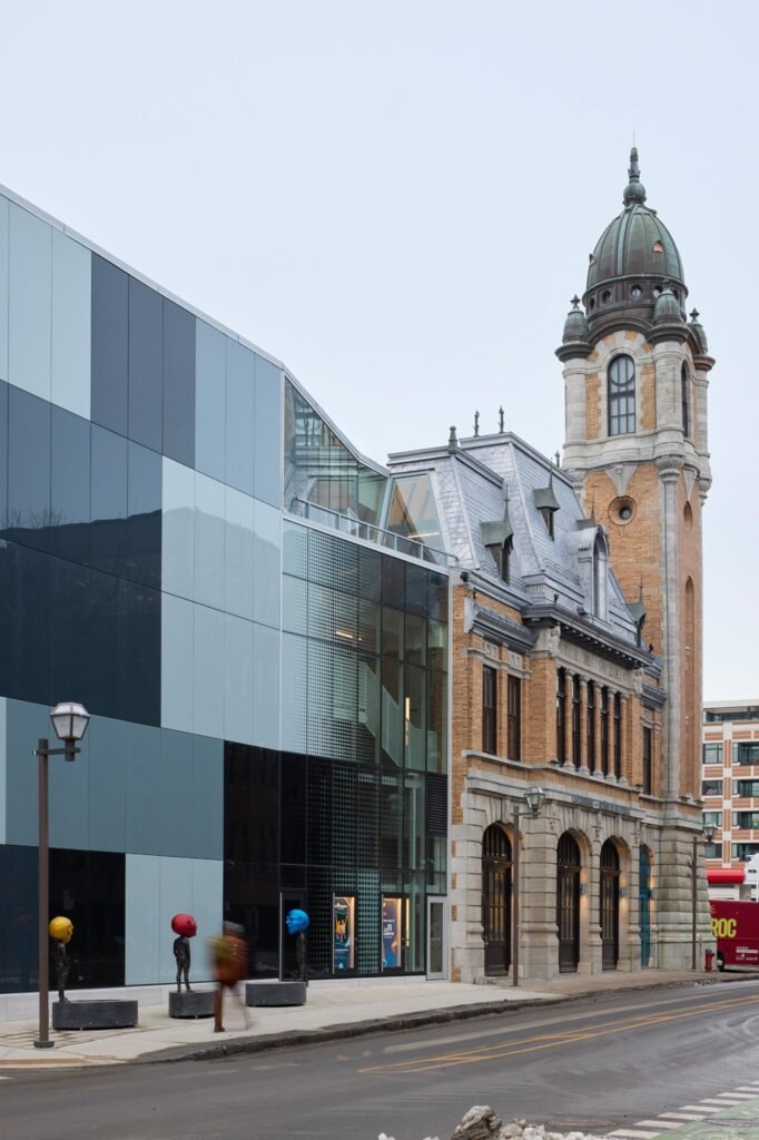 A vertical street-level view highlighting the stark material contrast between the crisp, geometric checkered glass facade of the new theatre addition on the left and the highly decorative brick and stone tower of the 1912 fire station on the right.