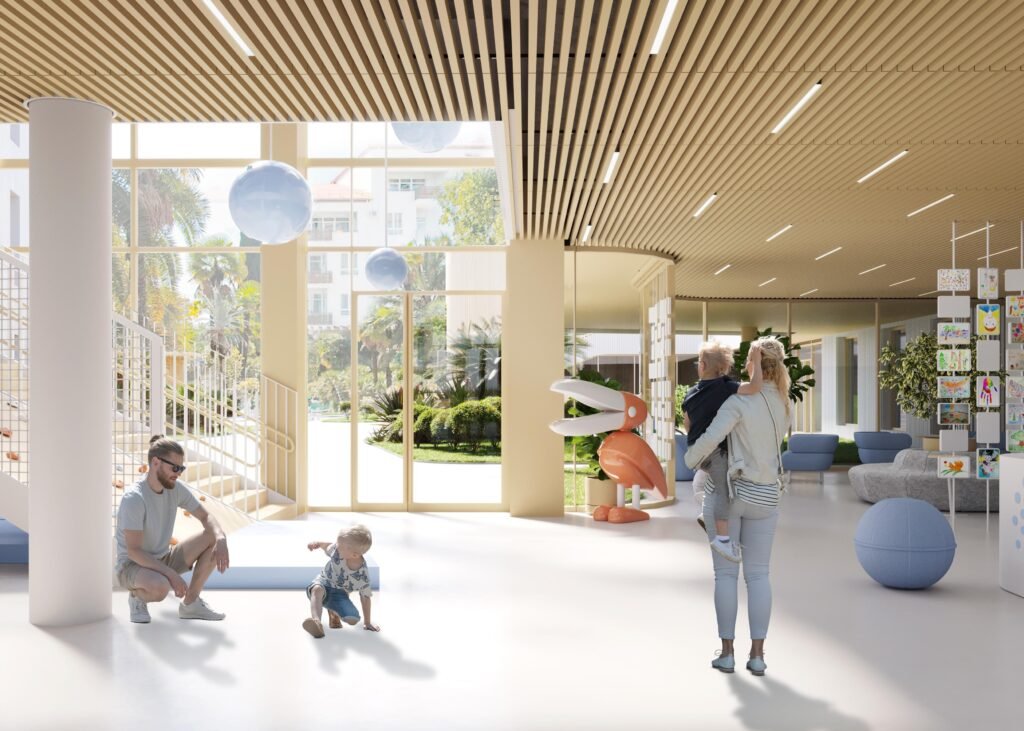 A bright, double-height indoor play area with floor-to-ceiling windows showing palm trees outside. A large, stylized orange pelican sculpture sits in the center. Parents and young children interact on a clean white floor with suspended blue spheres overhead, near a gallery display of colourful children's artwork.