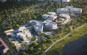 High-angle digital aerial rendering of the completed school campus. It reveals a series of interconnected, light-wood and white building blocks featuring intensive green roofs. The school borders a lush, green woodland and a natural wetland pond (ornithological park) with a system of walking trails.