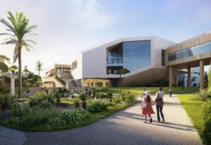 Exterior perspective rendering of the school complex approach. To the left is the public-facing congress centre wing featuring dramatic angular glazing. To the right, an enclosed bridge spans over a wide asphalt footpath. Landscaped lawns with shrubs, ornamental grass, and palm trees frame the walk.