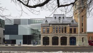 A wide exterior elevation showing the entire street facade of the complex on Dalhousie Street: the original 1912 brick fire station with its three arched wooden doors and bell tower is seamlessly combined with the new, geometric glass-and-steel addition on the left.