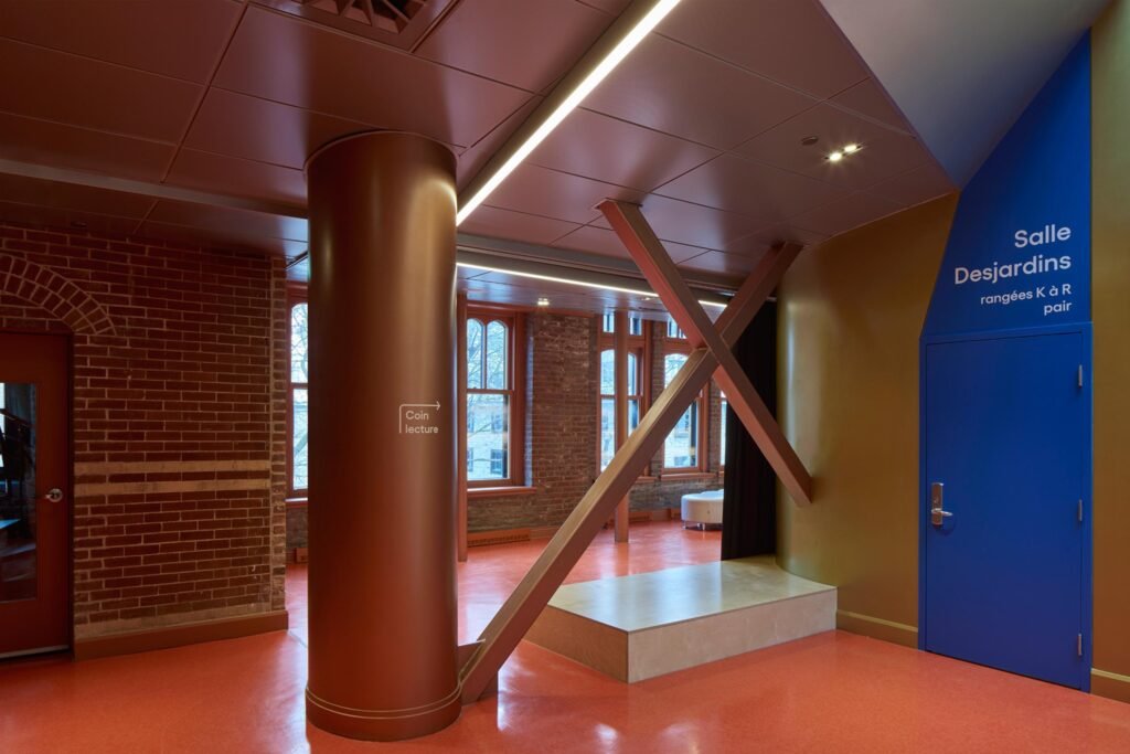 An interior view of the second-floor lobby area showing structural steel columns and cross-bracing clad in a metallic copper-orange finish, set against a red floor, green walls, and a bright blue door labeled "Salle Desjardins