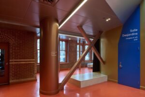An interior view of the second-floor lobby area showing structural steel columns and cross-bracing clad in a metallic copper-orange finish, set against a red floor, green walls, and a bright blue door labeled "Salle Desjardins