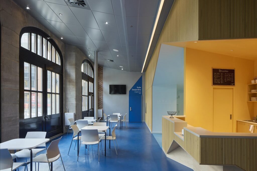 An interior view of the foyer dining area showing white modern tables and chairs on a deep blue floor, framed by the restored large arched black-painted wooden carriage doors of the fire station on the left, a bright blue door in the center, and a yellow service bar on the right