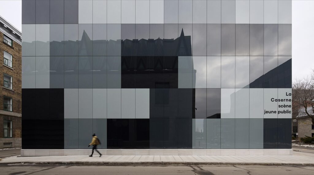 A flat, straight-on exterior elevation of the new theatre addition, demonstrating the geometric checkered layout of grey, black, and light glass panels that reflect a soft, overcast sky, with a pedestrian walking along the sidewalk below.