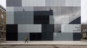 A flat, straight-on exterior elevation of the new theatre addition, demonstrating the geometric checkered layout of grey, black, and light glass panels that reflect a soft, overcast sky, with a pedestrian walking along the sidewalk below.