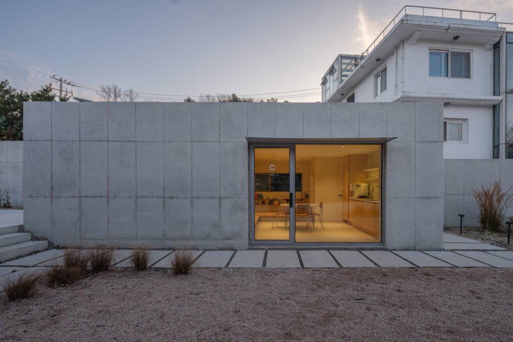 Exterior view of a separate concrete building with a large glass door revealing a warm kitchen and seating area inside.